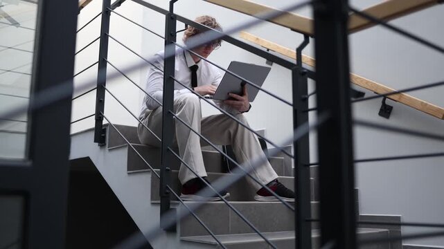 Office worker sitting on stairs working on laptop after hours staying focused and productive continuing tasks beyond workday showing dedication and overtime effort in workplace