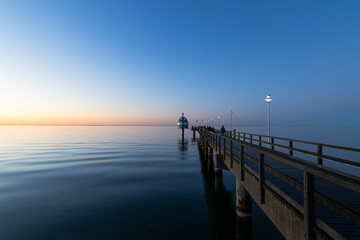 Fototapeta premium Seebrücke Zinnowitz bei ruhiger See zur Dämmerung mit Holzsteg und Lampen über der Ostsee unter klarem Himmel