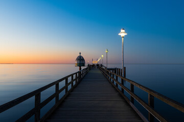 Fototapeta premium Seebrücke Zinnowitz bei ruhiger See zur Dämmerung mit Holzsteg und Lampen über der Ostsee unter klarem Himmel