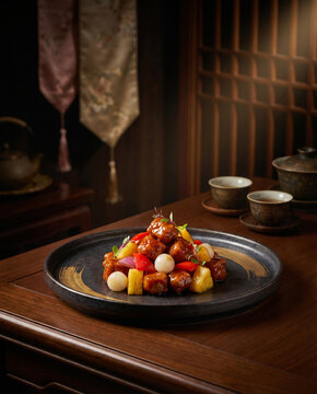 Plated sweet and sour chicken dish with pineapple chunks, white pearl onions, and red and yellow bell peppers on a dark ceramic plate on wooden table with traditional teacups and teapot in warm ambien