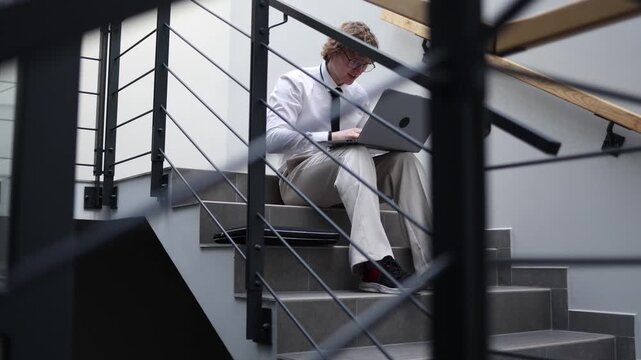 Focused worker sitting on office stairs working on laptop staying productive and determined during quiet downtime managing tasks and goals in modern workspace