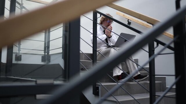 Stressed businessman sitting on office stairs working on laptop feeling pressure fatigue and burnout during workday struggling with deadlines and mental overload in modern workspace