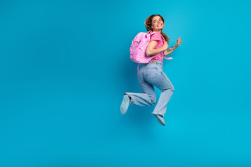 Naklejka premium Young woman with pink backpack jumps in the air against a blue background wearing striped tshirt and jeans
