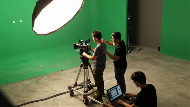 Professional cinematographers and crew operate a digital cinema camera with dolly in front of a large green screen, under bright studio lights.