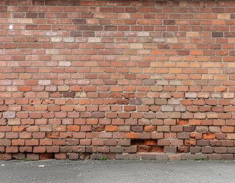 A weathered brick wall exhibits signs of age and erosion, with crumbling sections and a textured surface against a paved ground.