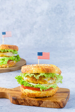A festive bagel burger styled for Independence Day in the USA. Two bagel sandwiches sit on a cutting board, filled with egg, lettuce, and tomato on a gray background. Small flags decorate the top.