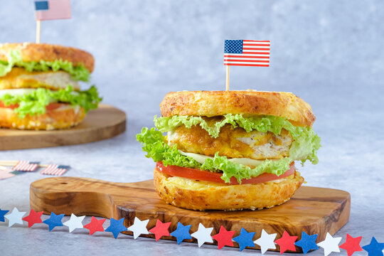 A festive bagel burger styled for Independence Day in the USA. Two bagel sandwiches on a cutting board, filled with crispy chicken, lettuce, and tomato on a gray background.
