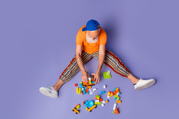 Obraz premium Older man with orange shirt and striped pants enjoys building with colorful blocks on a purple backdrop, a playful, carefree vibe that feels creative and hipster-inspired