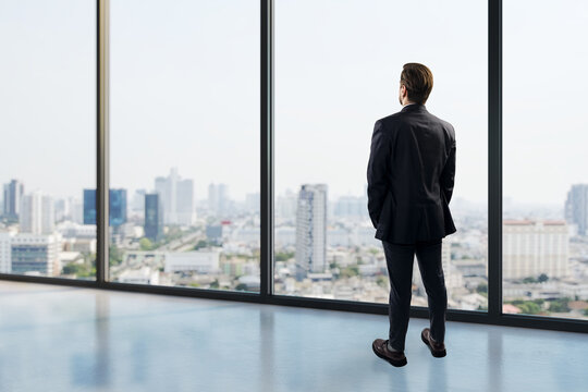Business person standing in modern office viewing urban city skyline through expansive large windows in bright interior background.