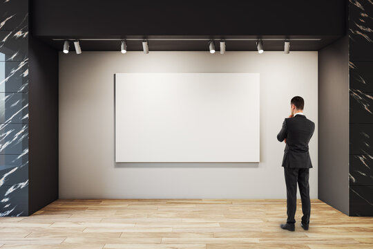 Blank gallery canvas observed by a lone businessman in a modern exhibition hall with spotlights illuminating a neutral background wall.