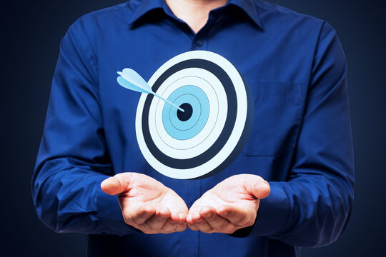 Business person holding floating target with arrow above open hands showing goal achievement concept on dark blue light background.