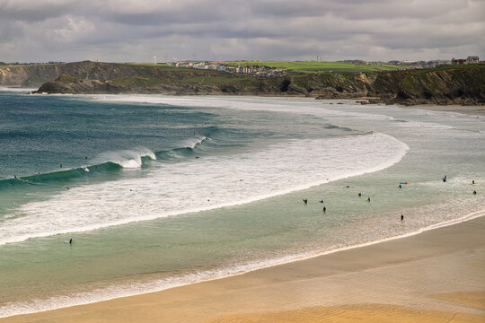 Ocean's rhythm surfers' patient dance. Surfers and bodyboarders enjoy the waves at a wide sandy beach backed by cliffs and a coastal town under an overcast sky.