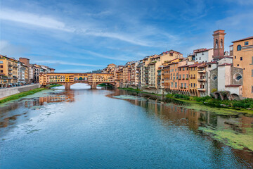 Fototapeta premium Ponte Vecchio bridge stands over the Arno River in Florence, surrounded by historic buildings. Boats glide beneath the bridge under a sanny sky.
