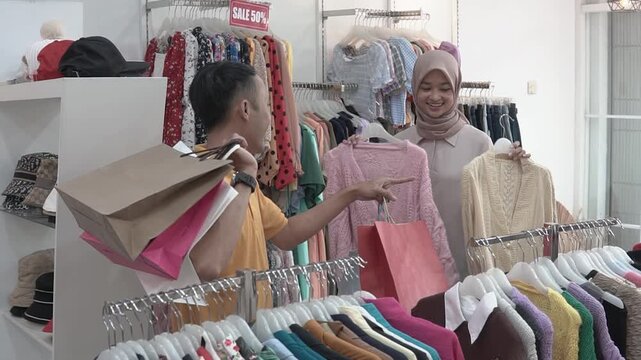 Asian couple comparing sweaters and bags at busy boutique, woman in hijab holds knit sweater while male shopper carries colorful