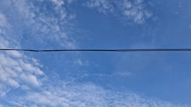 A clear blue sky background with soft scattered white clouds intersected by a single straight dark electrical wire crossing horizontally