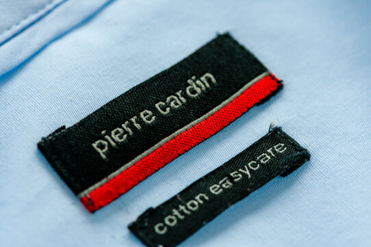 Macro shot of a black and red Pierre Cardin woven brand label stitched onto a light blue cotton men's shirt beside a smaller cotton easycare tag.