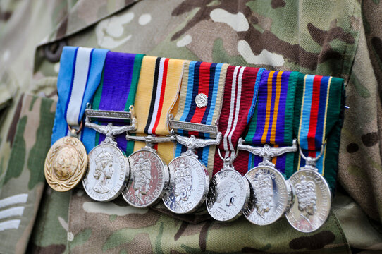 A British soldier in combat uniform stands with chest adorned with a row of service medals, including those for operations in Afghanistan and Kosovo, symbolizing a career of commitment and duty.