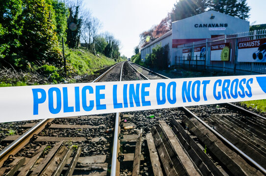 A close-up of blue and white police cordon tape stretched across a gravel-ballasted railway track, marking a secure crime scene investigation following a tragic fatal incident on the rails.