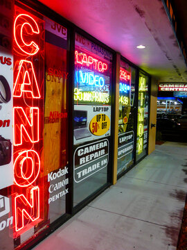 A vibrant evening street scene featuring the illuminated storefront of a camera retailer, dominated by bright neon brand logos for Canon and Nikon glowing against the urban night.
