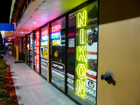 A vibrant evening street scene featuring the illuminated storefront of a camera retailer, dominated by bright neon brand logos for Canon and Nikon glowing against the urban night.