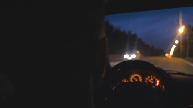 Close up of young guy holds hands on steering wheel and drives a modern automobile at evening. Male driver moves in his SUV car on empty road. Man rides in auto on highway at night time. Slow motion
