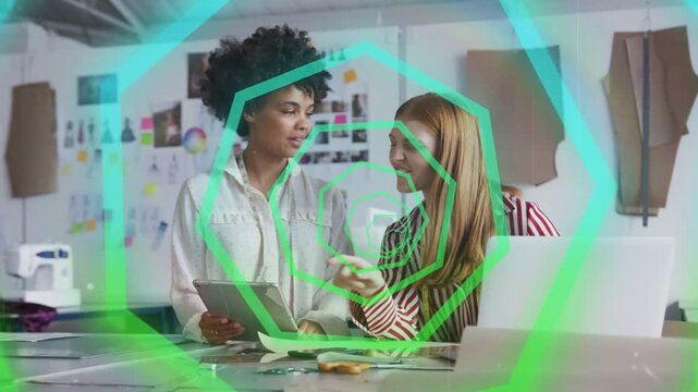 Two women pointing at tablet and collaborating on layout in design studio, hexagon overlay pulsing