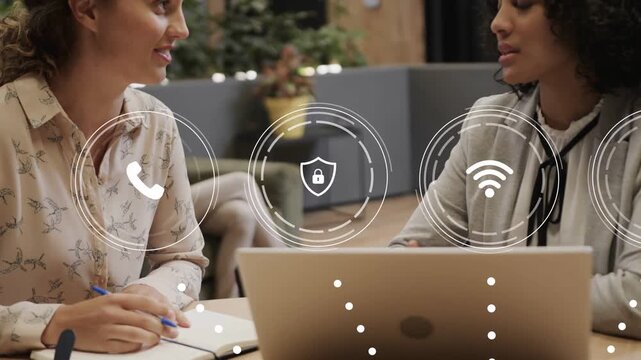 Left woman noting as right woman gesturing, laptop activating package icons for logistics planning