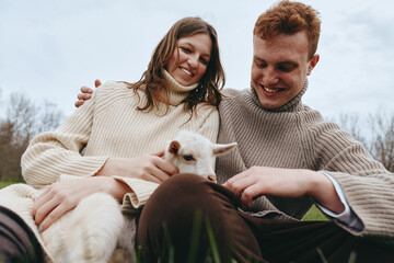 Naklejka premium A couple enjoys a playful moment with a friendly goat kid outdoors, wearing cozy sweaters, smiling warmly, capturing companionship and rustic lifestyle in a sunny countryside scene.