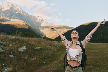 Naklejka premium Woman traveler with backpack raises arms in triumph in a meadow beneath mountains, enjoying fresh air and sunlight, embracing nature and outdoor adventure in a carefree lifestyle