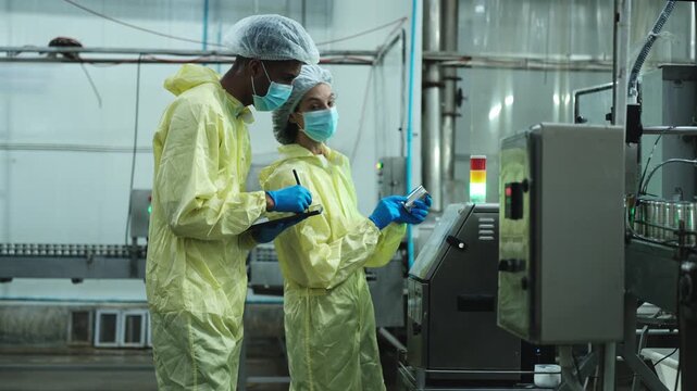 Two industrial engineer supervisors examine quality of steel containers on factory conveyor belt, inspect production process in canned food manufacture, and protective uniforms for hygiene operations.