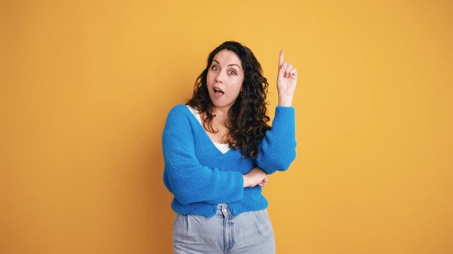 Pensive woman with curly hair thinking, then having a moment of insight and smiling with her finger up. Eureka moment on a yellow background