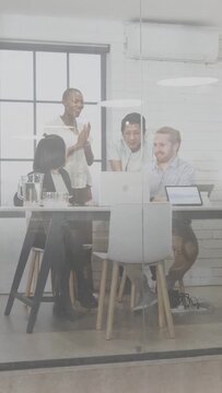 Vertical video: Seated woman gesturing at laptop, tech team leaning in, cheering win via glass wall
