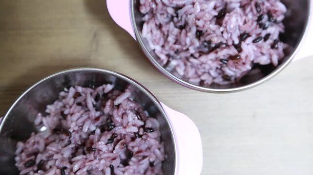 대한민국 서울의 가정집에서 아이들을 위해 준비된 두 개의 분홍색 식기에 건강한 흑미밥이 나란히 차려진 모습. 성장기 어린이를 위한 영양가 있는 식단과 일상적인 가정식 풍경을 따뜻하게 담아낸 하이 앵글 클로즈업 장면.