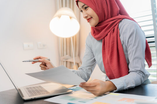 A Muslim businesswoman is working with financial graphs and a computer, remotely from a modern office.