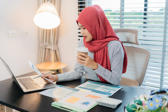 A Muslim businesswoman is working with financial graphs and a computer, remotely from a modern office.
