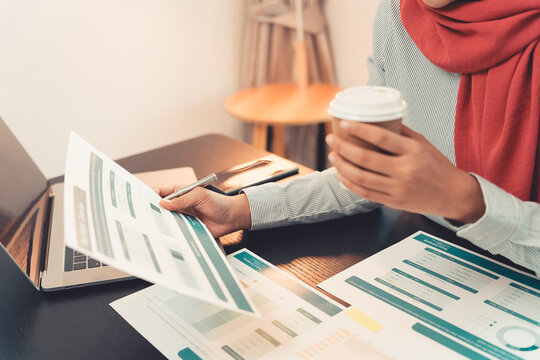 A Muslim businesswoman is working with financial graphs and a computer, remotely from a modern office.
