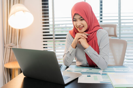 A Muslim businesswoman is working with financial graphs and a computer, remotely from a modern office.