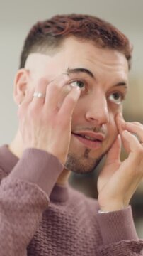 Handsome young gay man with a beard and piercing applying moisturizing face cream for his daily beauty and skincare routine at home