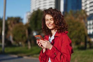 Fototapeta premium Smiling woman with curly hair wearing a red jacket checks her smartphone in a sunny park. Casual lifestyle, outdoor moment, bright mood, candid urban greenery and vitality.