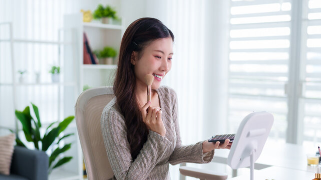 young Asian women applying makeup with brush watching tutorial on laptop computer learning beauty routine sitting at desk home office bright modern interior
