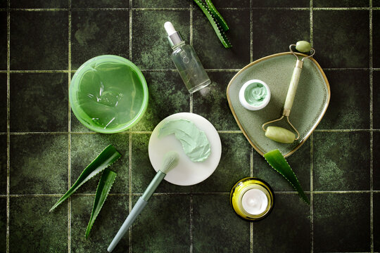 Flat lay of natural skincare products with aloe vera, serum, jade roller, and organic creams on dark green tiles. Concept of holistic beauty and wellness routine.