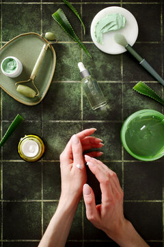 Vertical shot of hands applying white cream surrounded by aloe vera and natural cosmetics. Concept of hydration and hand care routine on an aesthetic green background.