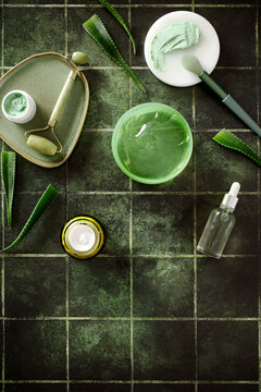 Top-aligned natural skincare products on dark green tiles with bottom copy space. Professional composition featuring serum, jade roller, and fresh aloe vera leaves.
