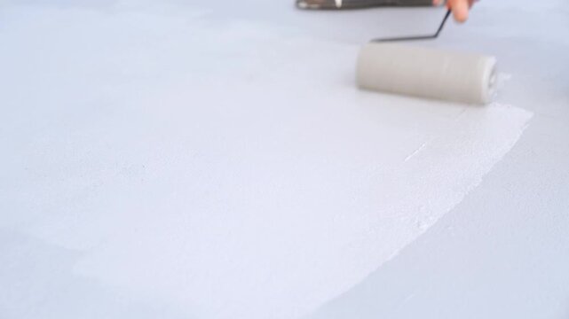 Hand using a roller to apply white liquid membrane for waterproofing on a flat roof surface. Motion blur effect showing active construction, industrial coating or floor renovation process.