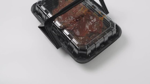 Slow-cooked barbecue ribs covered in thick sauce in a black plastic takeaway container on a white background. Ready-to-eat meal in disposable food packaging for delivery or takeout service.