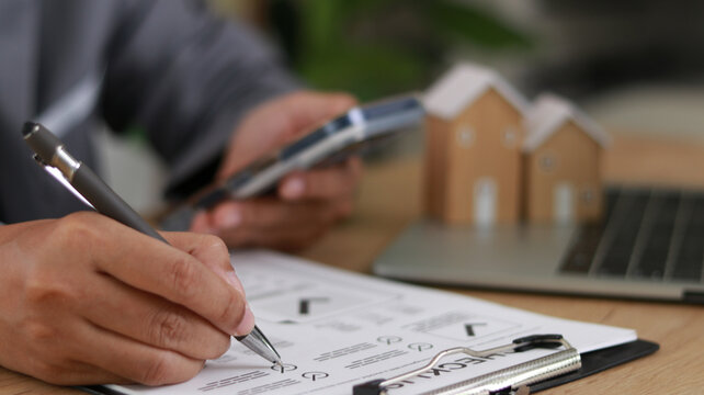 Close-up of professional reviewing documents and writing notes while using smartphone, with house models in background. Concept of real estate planning, mortgage, and property investment.