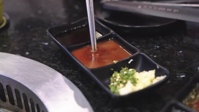 Metal chopsticks dipping into spicy red sauce in a black condiment tray. Includes minced garlic, green chili, and soy sauce. Traditional seasoning for Korean BBQ or Japanese yakiniku restaurant.