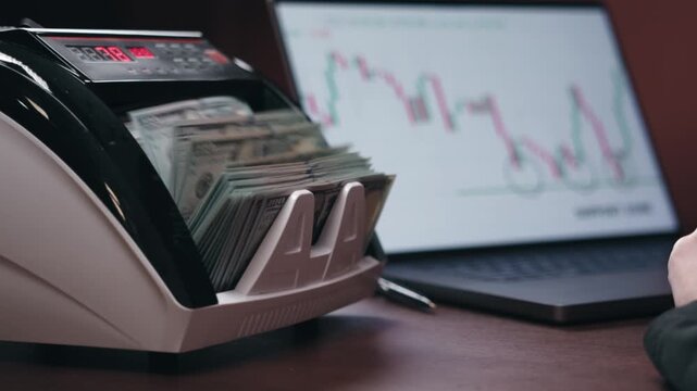Close-up of cash counting machine processing banknotes while a hand prepares to place more bills on a wooden desk with a laptop displaying financial graphs in the background