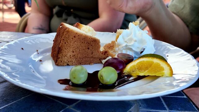 Close-up of a cheesecake slice served on a white plate with whipped cream, fresh grapes, orange slice and chocolate sauce, golden fork piercing the cake, natural lighting, hands visible.