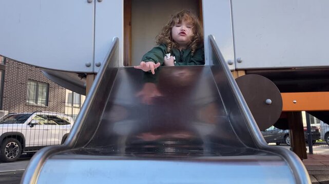 Curly hair child playing on slide at playground. Excited kid preparing for descent on metal chute. Active toddler enjoying outdoor game in public park setting.